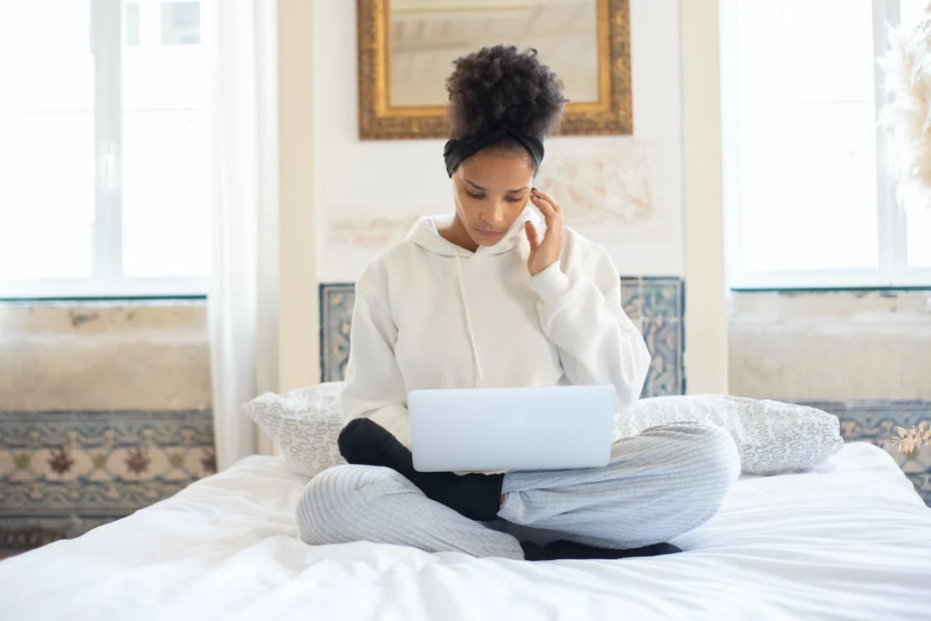 moça negra sentada na cama mexendo no notebook enquanto trabalha home office